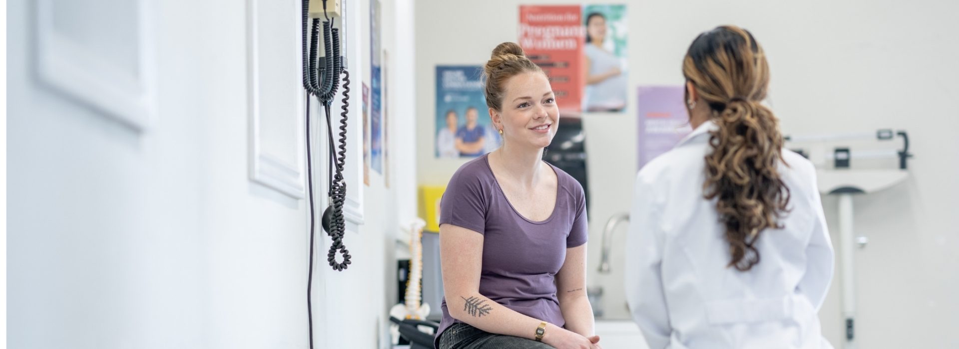A woman sits on an exam table, smiling and talking with a healthcare professional in a white coat inside a medical office with health posters on the wall.